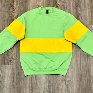 May include: A lime green crewneck sweatshirt featuring a wide, horizontal yellow stripe across the chest and upper sleeves. The garment has long sleeves, a ribbed collar, and ribbed cuffs, with a black brand label visible at the inner neckline.