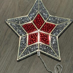 May include: A five-pointed star ornament featuring a central red geometric pattern surrounded by clear, textured glass-like segments. The star is framed in a gold-toned metal border and includes an attached white electrical cord with a plug.
