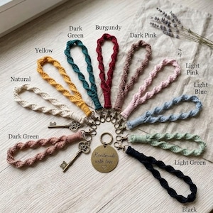 Op de afbeelding: Een set van tien macramé polsband-sleutelhangers in naturel, geel, donkergroen, bordeaux, donkerroze, lichtroze, lichtblauw, lichtgroen en zwart. Elke hanger heeft een metalen karabijnhaak, twee zijn bevestigd aan antieke sleutels en één aan een ronde messing hanger met de tekst Handmade with love.