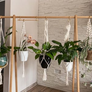 Può includere: Un supporto in legno chiaro a forma di A con una barra orizzontale e cinque portapiante in macramè sospesi tramite anelli dorati. I supporti contengono vari vasi, tra cui un vaso color ottanio, un vaso bianco con orchidee, un vaso nero, un vaso bianco e un vaso marrone testurizzato.