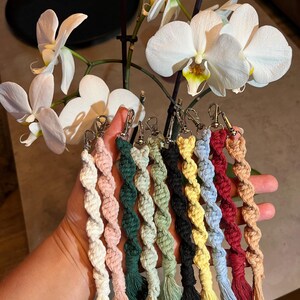 May include: A collection of nine macrame keychains in various colours including white, pink, dark green, light green, black, yellow, light blue, red, and tan. Each keychain features a twisted cord design, a silver metal lobster clasp, and a tassel.