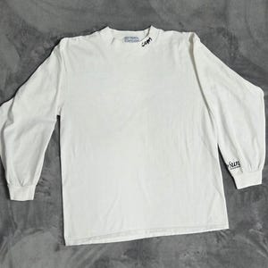Peut inclure: Un t-shirt blanc à manches longues avec un col rond. Le vêtement présente une inscription noire sur le col indiquant .com et une inscription noire sur le poignet de la manche gauche indiquant The Sun. Le devant est uni et sans motif.