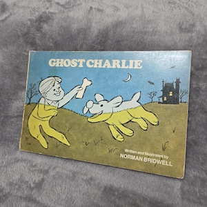 May include: A rectangular children's book titled Ghost Charlie, written and illustrated by Norman Bridwell. The cover features a cartoon illustration of a child in yellow clothing holding a bone for a white dog, set against a blue sky and a dark house.