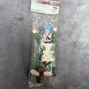 May include: A wooden nutcracker ornament in a clear plastic bag with a header card labeled SANTA'S WORLD DECORATION. The nutcracker has a light blue hat, green arms and legs, a yellow and green jacket, and a black mustache, with white hands and feet.