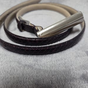 May include: A thin belt featuring a dark burgundy snakeskin-textured strap and a polished silver-tone metal bar buckle. The belt is coiled in a circular shape, revealing a light beige interior lining and a matching dark burgundy belt loop.