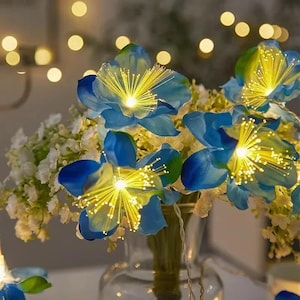 May include: A bouquet of artificial blue flowers with bright yellow fiber optic centers that emit a warm glow. The arrangement includes small white filler flowers and is displayed in a clear glass vase, creating a luminous display of light.