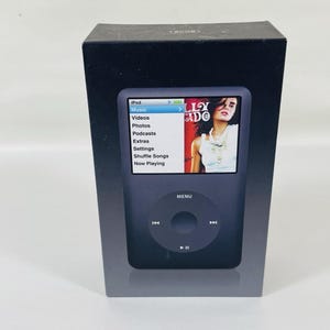 May include: A black rectangular product box featuring a graphic of a digital music player on the front. The screen displays a menu with options for Music, Videos, Photos, Podcasts, Extras, Settings, Shuffle Songs, and Now Playing.