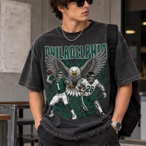 Può includere: Una t-shirt grigio antracite con lavaggio acido caratterizzata da una stampa grafica con il testo PHILADELPHIA in verde. Il design include una grande aquila con le ali spiegate sopra due giocatori di football scheletrici in divise verde e bianco.