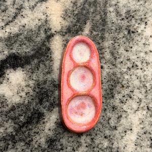May include: An oval-shaped ceramic tray featuring three circular indentations. The exterior rim is a vibrant coral-pink, while the interior of each circular well transitions into a lighter, iridescent white and pink speckled glaze finish.