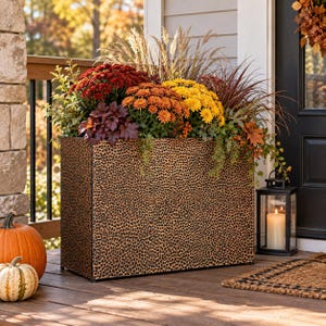May include: A rectangular planter with a black and tan leopard print pattern, filled with a vibrant autumn arrangement of red, orange, and yellow chrysanthemums, purple foliage, and tall ornamental grasses, positioned on a wooden porch.