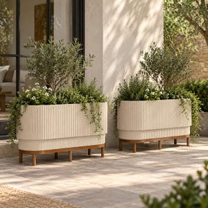 May include: Two elongated, off-white planters with a vertical ribbed texture, each supported by four wooden legs. The planters are filled with green olive trees, small white flowers, and trailing vines, creating a layered botanical display.