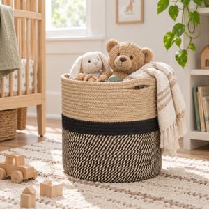 May include: A round woven rope storage basket featuring a two-tone design with a natural tan upper section and a black and tan patterned lower section. The basket contains a plush teddy bear, a plush rabbit, and a cream-colored fringed throw blanket.