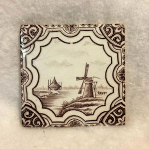 May include: A square ceramic tile featuring a sepia-toned illustration of a Dutch landscape with a windmill and a small boat on the water. The tile has a decorative border with floral and scroll patterns and a visible crackled glaze finish.