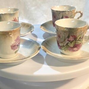 May include: A set of four porcelain teacups and matching saucers featuring a delicate floral pattern with pink roses and green leaves. Each cup and saucer is finished with a gold-toned rim and handle, showcasing a classic design with white interiors.