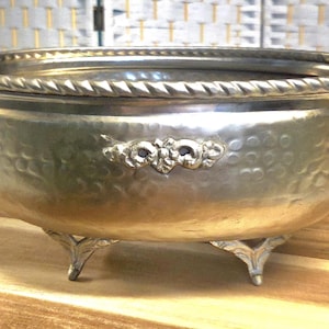 May include: A silver-toned metal bowl with a hammered texture, featuring a rope-style rim, two side handles, and three decorative feet. The exterior surface is adorned with a central embossed floral motif on both the front and back sides.