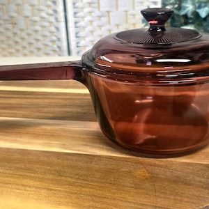 May include: A translucent amber-colored glass saucepan with a matching lid. The pot features a long, flat handle extending from the side, and the circular lid has a central knob with a radiating sunburst pattern etched into the glass surface.