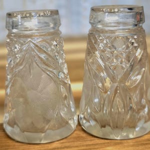May include: A pair of clear glass salt and pepper shakers featuring a pressed pattern of geometric diamonds and a central floral motif. The shakers have a tapered cylindrical shape with a smooth rim at the top and a textured base design.