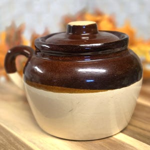 May include: A two-toned ceramic crock with a lid and a side handle. The upper portion and lid feature a glossy dark brown glaze, while the lower body is finished in a smooth, matte cream color. The vessel has a rounded shape and a small knob.