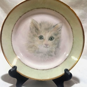 May include: A round decorative plate featuring a central illustration of a fluffy kitten with light fur and green eyes. The plate has a pale pink center, a wide light green border, and a thin gold rim. It is displayed on a black plastic stand.