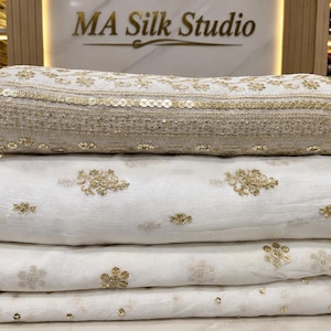 Könnte beinhalten: Ein Stapel aus vier gefalteten weißen Stoffstücken mit goldener Stickerei und Pailletten. Das oberste Stück zeigt ein dichtes horizontales Band aus goldenen Blumenmustern und Pailletten, während die unteren Stücke verstreute Blumenmotive aufweisen.