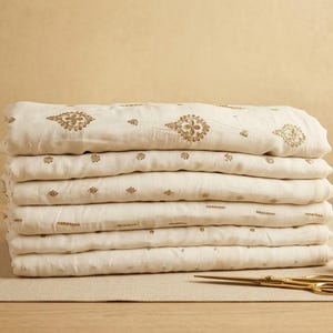 Könnte beinhalten: Ein Stapel gefalteter cremeweißer Stoff mit goldfarbenen, gestickten Blumenmotiven. Neben dem Stoff liegen eine Spule mit goldenem Garn und eine goldfarbene Schere. Das Textil zeigt ein wiederkehrendes Muster aus kleinen, filigranen Golddesigns.