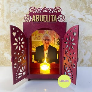 Puede incluir: Un altar de madera de color rojo oscuro con dos puertas abiertas que presentan intrincados recortes florales. La parte superior tiene la palabra ABUELITA en letras doradas. Dentro, una foto de una persona mayor sobre una vela LED.