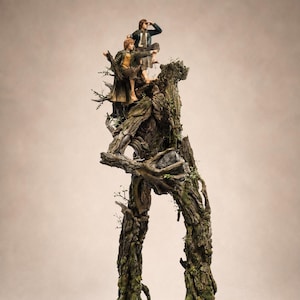 Op de afbeelding: Een gedetailleerd beeldje met twee kleine figuren die bovenop een groot, antropomorf boomwezen staan. De boom heeft een getextureerde, schorsachtige huid in bruine en groene tinten, met mosachtige accenten. De figuren dragen kleding in aardetinten.