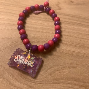 Puede incluir: Una pulsera de cuentas con esferas alternas en color morado y rosa ensartadas en un cordón. Un dije rectangular de color morado translúcido con destellos iridiscentes y la palabra "Sweet" escrita en blanco cuelga de una argolla metálica.
