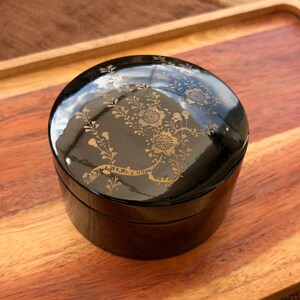 May include: A round black lacquer container with a glossy finish, featuring a delicate gold-colored floral design on the lid. The cylindrical box has a smooth, reflective surface and a matching lid that sits flush with the base of the container.