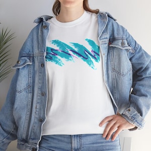 May include: A white crew neck t-shirt featuring a graphic print of teal and purple brushstroke waves across the chest. The t-shirt is worn underneath a light blue denim jacket with metal buttons and a classic collar, paired with blue denim jeans.
