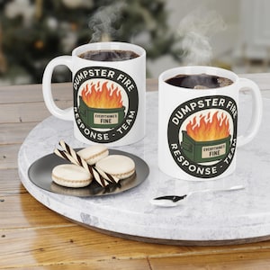 May include: Two white ceramic mugs filled with dark liquid, each featuring a circular graphic of a green dumpster on fire with the text "DUMPSTER FIRE RESPONSE TEAM" and "EVERYTHING'S FINE" on the side, accompanied by a small plate of biscuits.