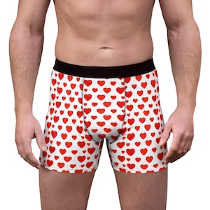 May include: Boxer briefs featuring a repeating pattern of small red hearts on a white background. The garment includes a solid black elastic waistband and visible black stitching along the front centre panel and leg seams for structure.