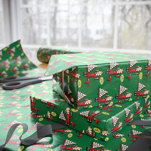 May include: Green wrapping paper featuring a repeating cartoon pattern of an elf in a red suit holding a stack of white gifts. The paper is used to wrap rectangular boxes, with a roll of the same paper and black scissors visible nearby.