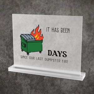 Op de afbeelding: Een doorzichtig acrylbord met een rechthoekige voet, voorzien van een afbeelding van een groene afvalcontainer die in brand staat. De tekst luidt: IT HAS BEEN DAYS SINCE OUR LAST DUMPSTER FIRE. Het bord is ontworpen voor op een tafel.