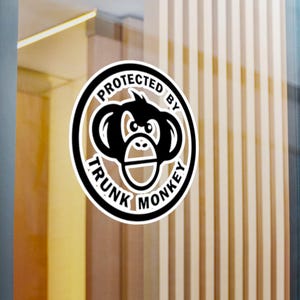 May include: A circular sticker featuring a black and white graphic of a monkey face centered within a thick border. The text "PROTECTED BY TRUNK MONKEY" is printed in bold black capital letters along the top and bottom of the circular frame.