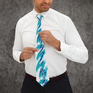 May include: A white necktie featuring a repeating pattern of teal brushstrokes and thin, jagged purple lines. The tie is worn with a crisp white long-sleeved button-down shirt and dark trousers, creating a sharp contrast against the patterned fabric.