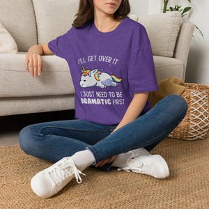Pode incluir: Uma t-shirt roxa de manga curta com o gráfico de um unicórnio deitado com crina e cauda arco-íris. O texto diz: I'LL GET OVER IT I JUST NEED TO BE DRAMATIC FIRST. A t-shirt é combinada com calças de ganga azuis e sapatilhas brancas.