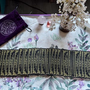May include: A deck of black tarot cards with gold foil celestial designs featuring suns and geometric patterns, arranged in a fan shape. Beside the cards is a purple velvet drawstring pouch with a white circular emblem and a crystal tree.
