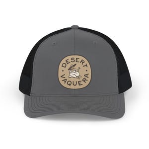 Op de afbeelding: Een antracietgrijze truckerpet met een zwarte mesh achterkant en een ronde patch op de voorkant. De patch heeft een beige achtergrond met donkerbruine tekst DESERT VAQUERA en een centrale illustratie van een persoon met een hoed op.
