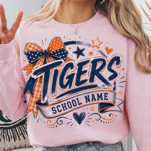 May include: A light pink sweatshirt featuring a graphic design with the word TIGERS in large navy blue brush-stroke lettering. Below, a banner reads SCHOOL NAME. The design includes a large orange and navy polka dot bow, stars, and hearts.