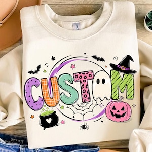 May include: A cream-coloured crewneck sweatshirt featuring a graphic print that spells the word CUSTOM. The letters are decorated with various patterns including purple dots, orange plaid, green stripes, and pink leopard print, alongside a ghost, a black witch hat, a jack-o'-lantern, a spider web, bats, and a cauldron.