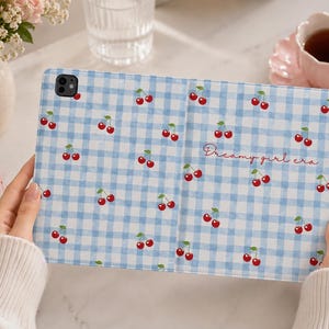 Peut inclure: Une housse de tablette à motif vichy bleu clair et blanc avec un imprimé de cerises répétitif et le texte cursif "Dreamy girl era" en rouge. L'étui présente une découpe noire pour l'appareil photo dans le coin supérieur gauche et un design pliable.