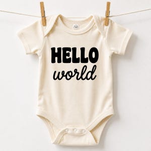 May include: A cream-coloured short-sleeve bodysuit featuring the text HELLO world in bold black lettering on the front. The garment has a round neckline, short sleeves, and three snap closures at the bottom, hanging from a clothesline.