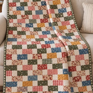 May include: A rectangular quilt featuring a patchwork pattern of small, alternating rectangular fabric blocks in various floral, plaid, and solid prints. The colours include red, blue, yellow, green, and white, finished with a dark green border.