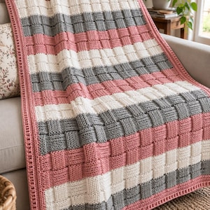 May include: A rectangular knitted throw blanket featuring a basketweave texture. The design consists of alternating horizontal stripes in dusty rose, cream, and charcoal gray. The blanket is finished with a solid dusty rose crocheted border.