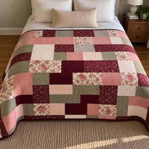 Puede incluir: Una colcha de patchwork con un patrón de bloques rectangulares en tonos burdeos intenso, rosa suave, verde salvia y crema. El diseño incorpora bloques de tela de colores sólidos junto a cuadrados con estampados florales y detalles botánicos.