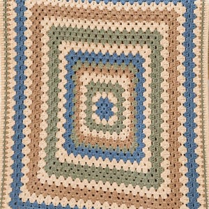 May include: A rectangular crochet blanket featuring a classic granny square pattern with concentric borders in alternating shades of cream, sage green, tan, and dusty blue. The textured yarn work creates a repeating geometric design throughout.