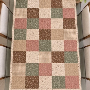 Pode incluir: Um caminho de mesa acolchoado retangular com um design de patchwork de quadrados alternados em tons de verde sálvia, rosa antigo, creme e castanho. Os quadrados de tecido apresentam estampados florais e padrões axadrezados.