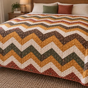 May include: A quilted bedspread featuring a repeating chevron pattern in alternating bands of cream, mustard yellow, dark brown, and burnt orange. Each colored stripe displays a small, intricate floral print, and the entire quilt is stitched with a grid pattern.