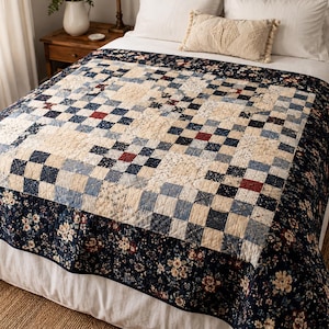 Könnte beinhalten: Eine Patchwork-Tagesdecke mit einem zentralen Gitter aus cremefarbenen, marineblauen und roten Quadraten, umgeben von einem breiten Rand aus dunkelblauem Stoff, der mit einem Blumenmuster aus kleinen weißen und roten Blüten sowie grünen Blättern verziert ist.